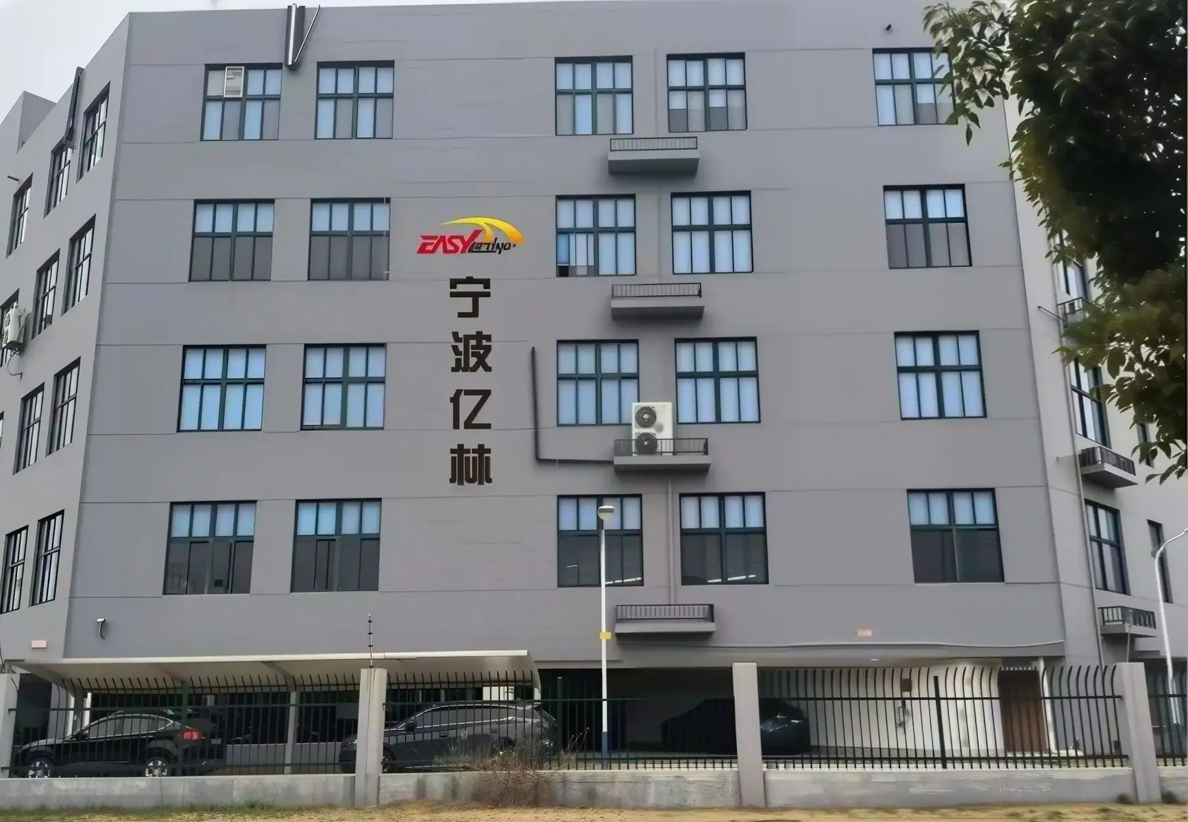 Modern multi-story building with Easy Lifting signage, featuring multiple windows, air conditioning units, and a parking area with cars, located in an urban setting.